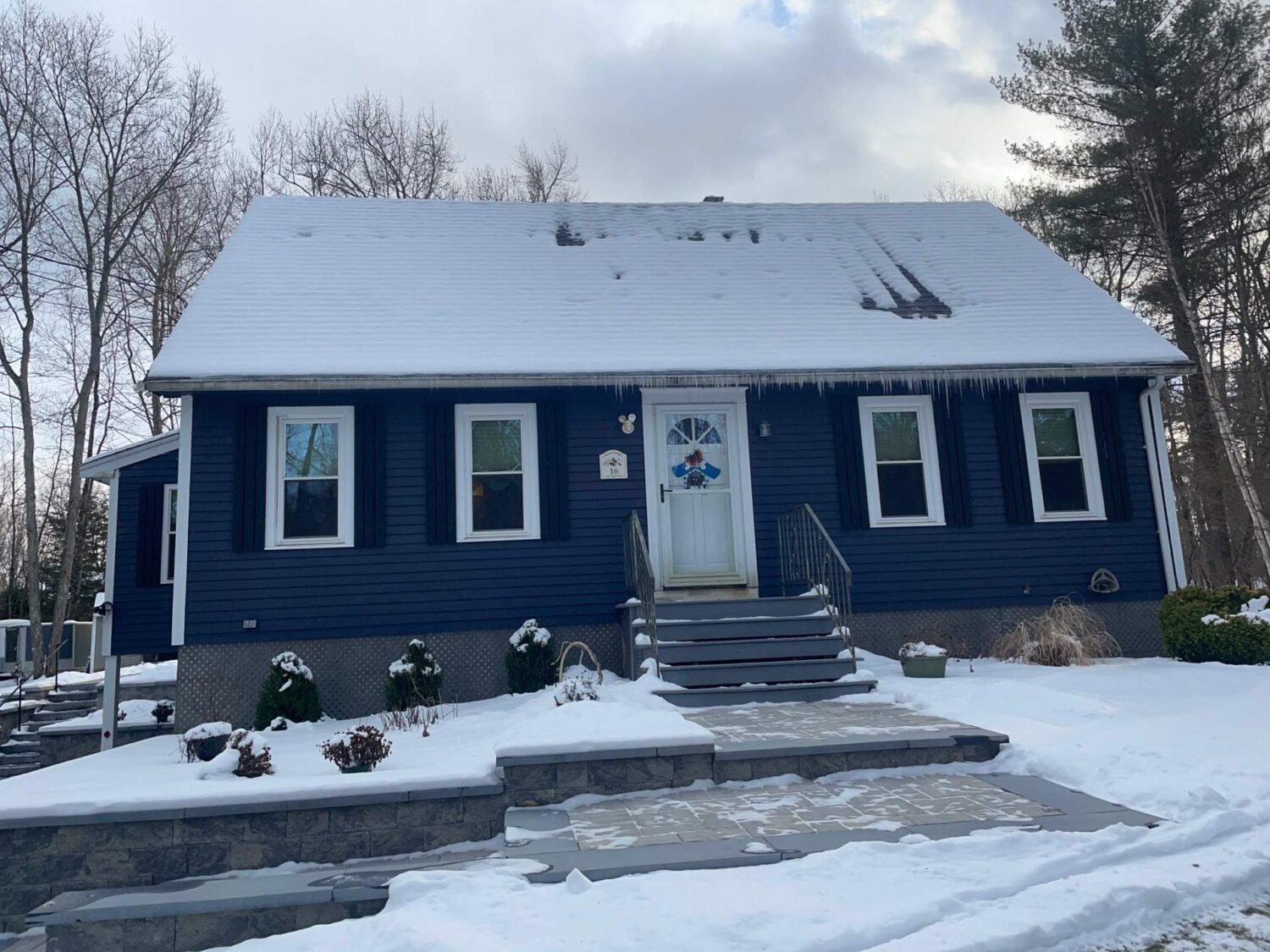 A blue house with white trim is covered in snow. Icicles hang from the roof, and patches of snow cover the steps and yard. Bare trees surround the house, and the sky is mostly cloudy.