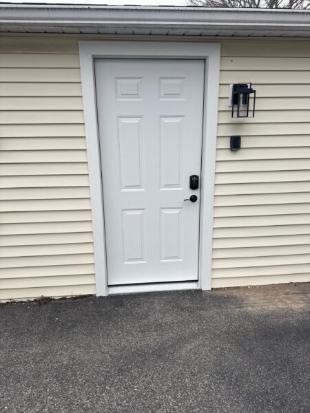 A closed white door with a keypad lock is set in light beige siding. A black wall-mounted light fixture is to the right of the door, above a patch of bare ground and asphalt driveway.
