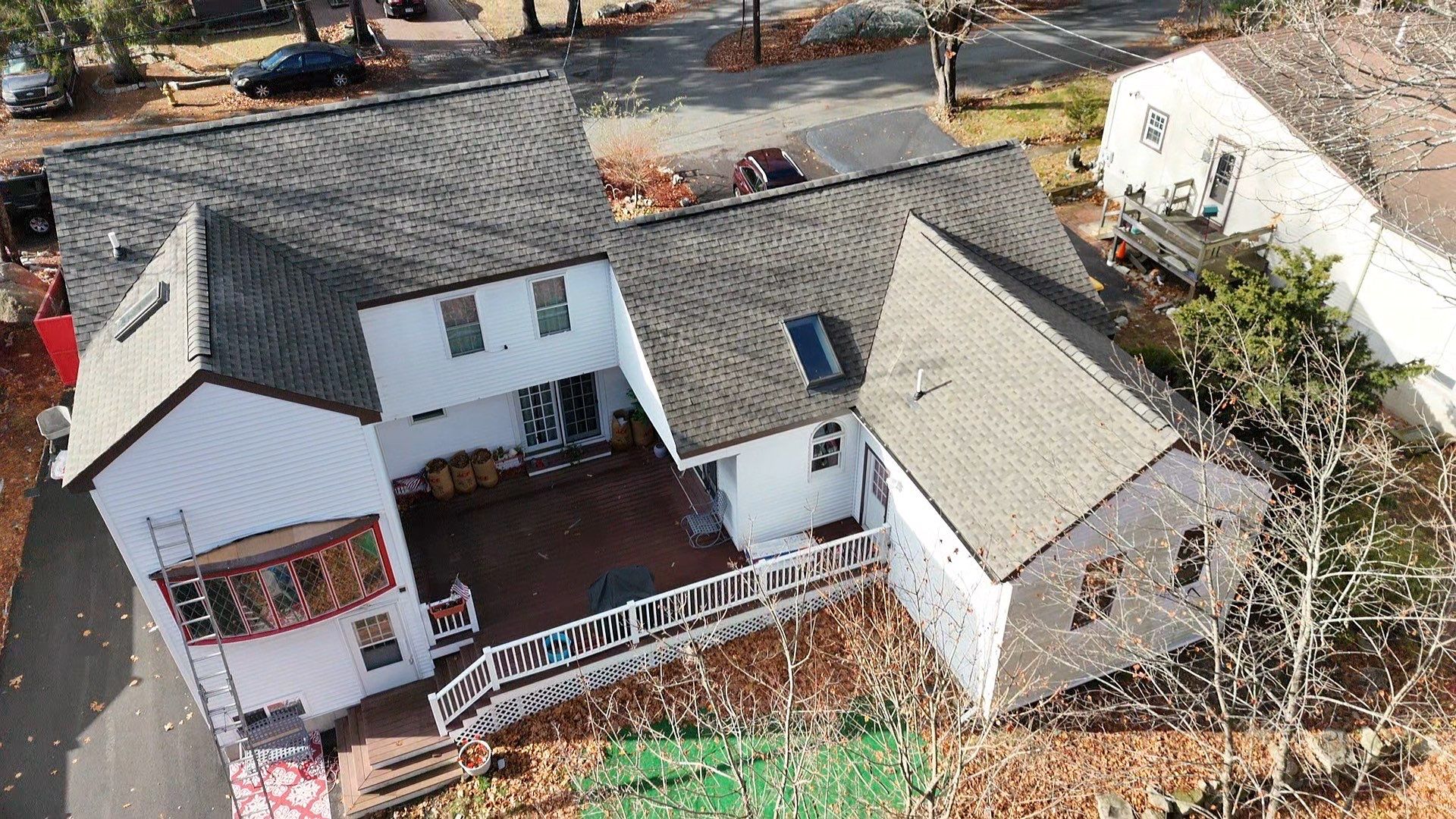 Aerial view of a white two-story house with a gray shingled roof, large wooden deck, white railing, and driveway. Fallen leaves are scattered around the yard—perfect for showcasing reliable emergency roof repair services.
