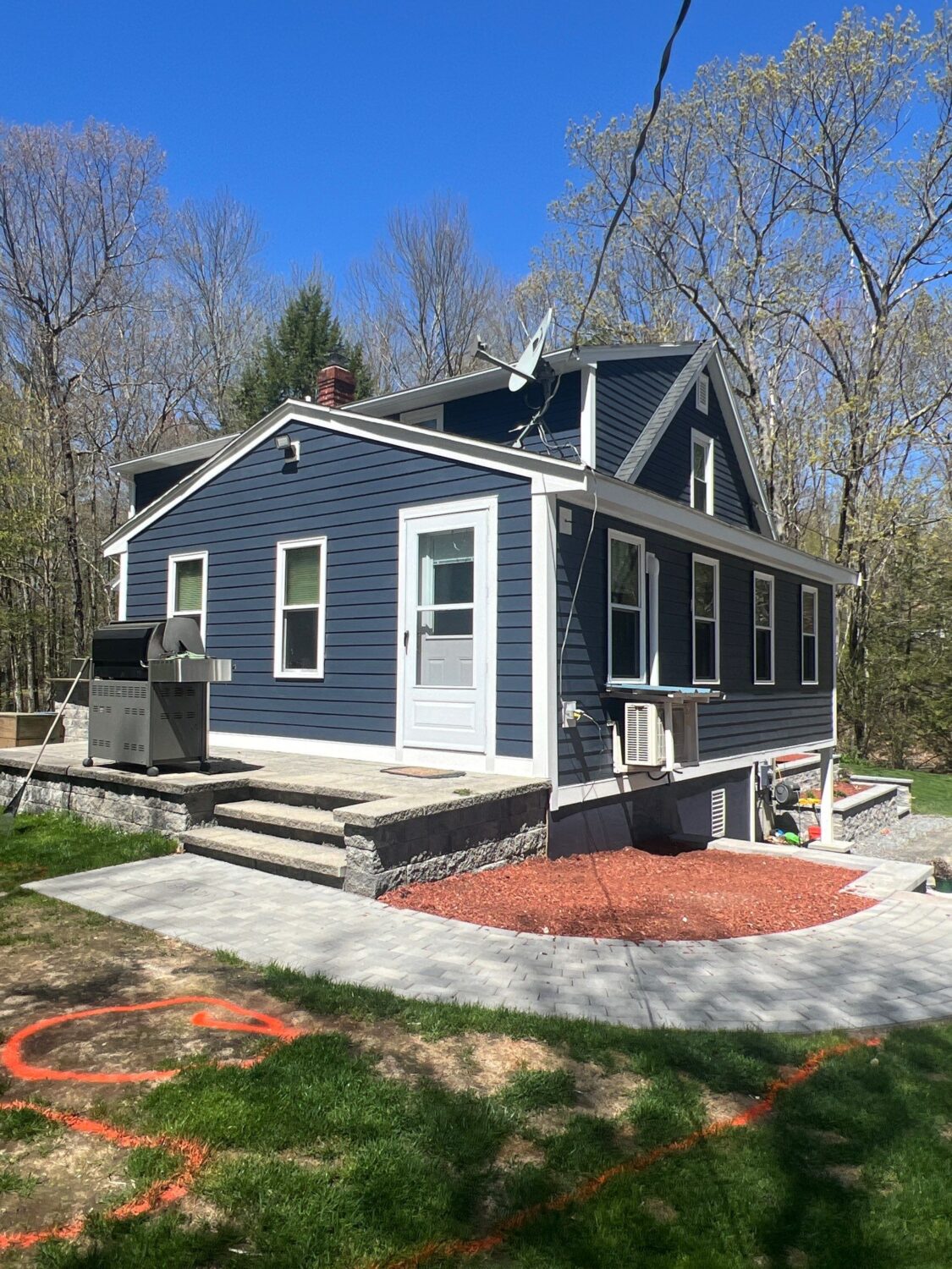 A small, blue house with white trim sits on a stone foundation, surrounded by trees and a clear blue sky. A barbecue grill is on the side patio, and orange spray paint marks are visible on the grass in the foreground.
