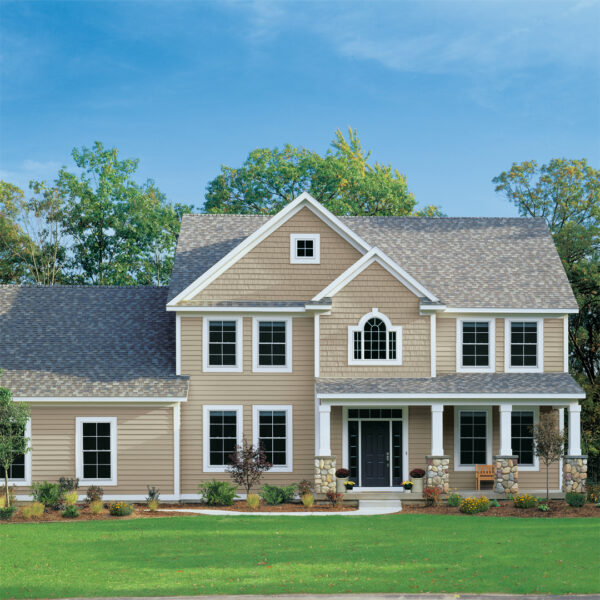 A two-story beige house with white trim, a covered front porch, black front door, and well-kept green lawn stands surrounded by trees under a blue sky—an example of work by the best Norwood home improvement companies.