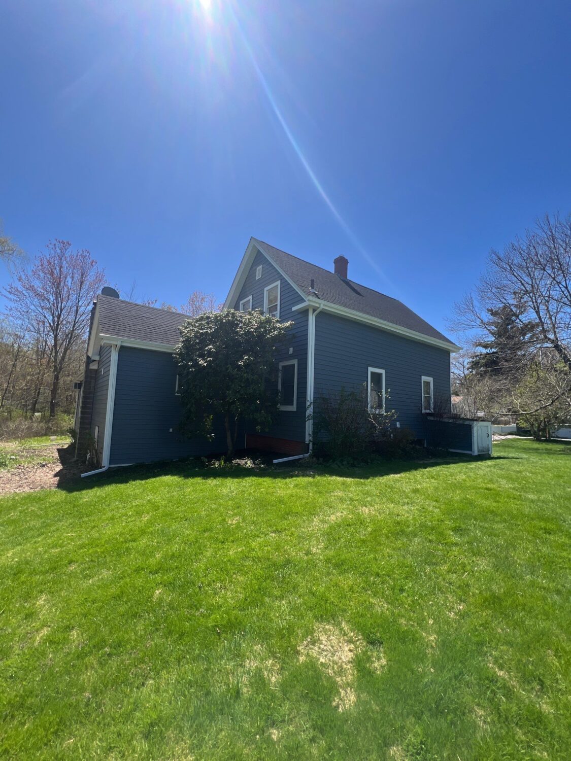 A dark blue house with white trim sits on a green lawn under a clear blue sky, with sunlight streaming down and trees with sparse leaves surrounding the yard.