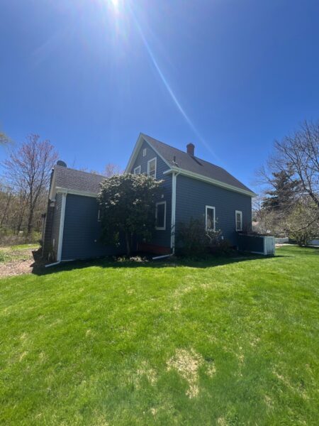 A dark blue house with white trim sits on a green lawn under a clear blue sky, with sunlight streaming down and trees with sparse leaves surrounding the yard.