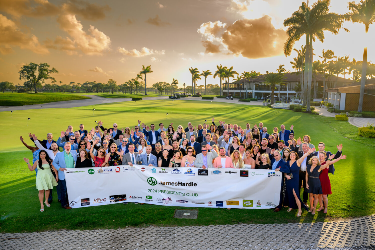 A large group of people in business and formal attire stand outdoors on a golf course, smiling and waving, holding a “James Hardie 2024 President’s Club” banner. Palm trees and a clubhouse are in the background, featuring top names like Coastal Windows & Exteriors.