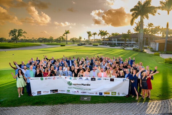 A large group in semi-formal attire stands on a green lawn, smiling and waving behind a banner that reads 2024 PRESIDENTS CLUB. Palm trees and a clubhouse are visible as Holliston Town News celebrates Coastal Windows & Exteriors 15th anniversary.