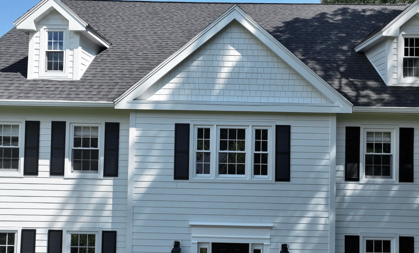 White two-story house with black shutters, a gray shingled roof, and several windows; shadows from trees are cast across the front of the house.