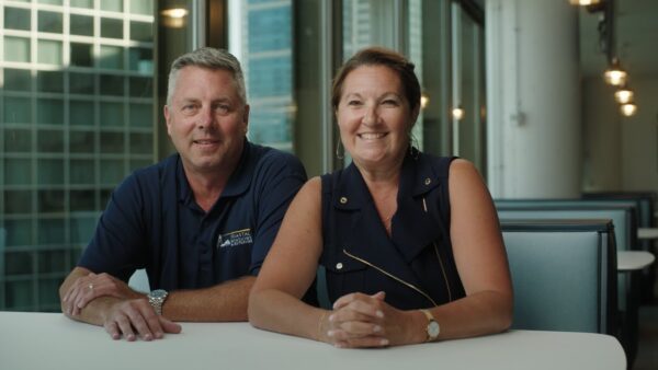 A man and a woman sit side by side at a white table in a modern office with large windows overlooking city buildings, smiling at the camera—professionals from one of the best Norfolk home improvement companies.