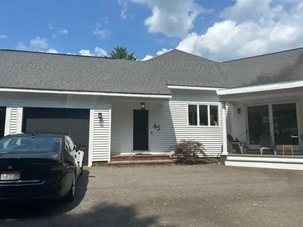 A white house with a black front door and the number 45, a black car in the driveway, and a porch with chairs on the right side under a partly cloudy sky—serviced by a trusted West Newton MA Roofing contractor.