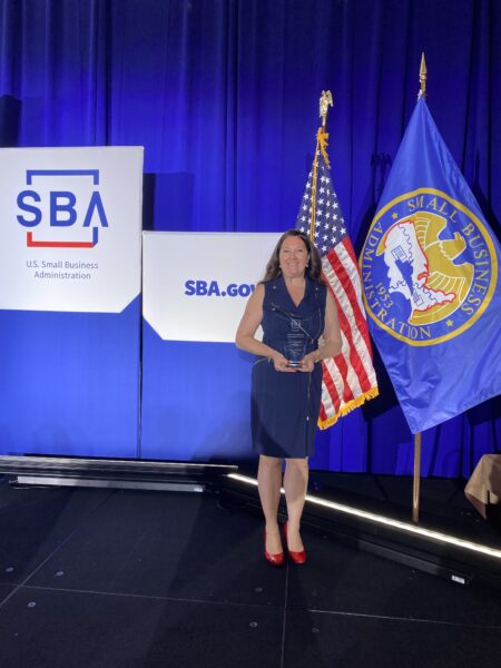A woman in a navy dress and red shoes stands holding an award on stage between the U.S. flag and the Small Business Administration flag, representing one of the best Natick home improvement companies, with SBA banners in the background.