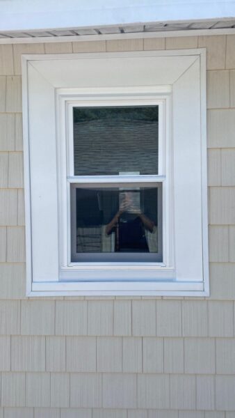 A double-pane window with a white frame set in beige siding. The reflection in the glass shows a person taking a photo, as well as the roof and siding of a nearby building.