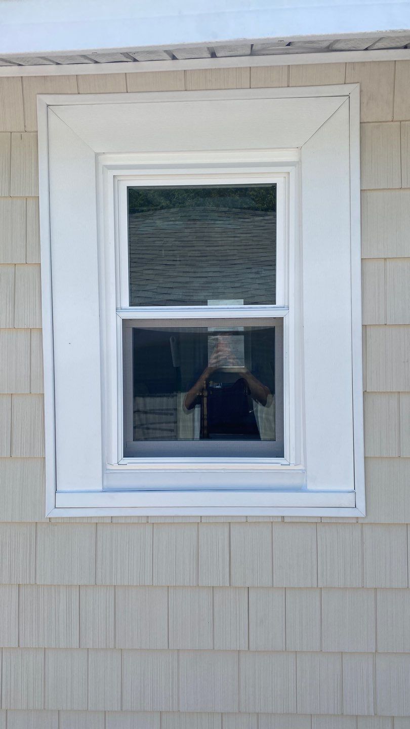 A double-pane window with a white frame set in beige siding. The reflection in the glass shows a person taking a photo, as well as the roof and siding of a nearby building.