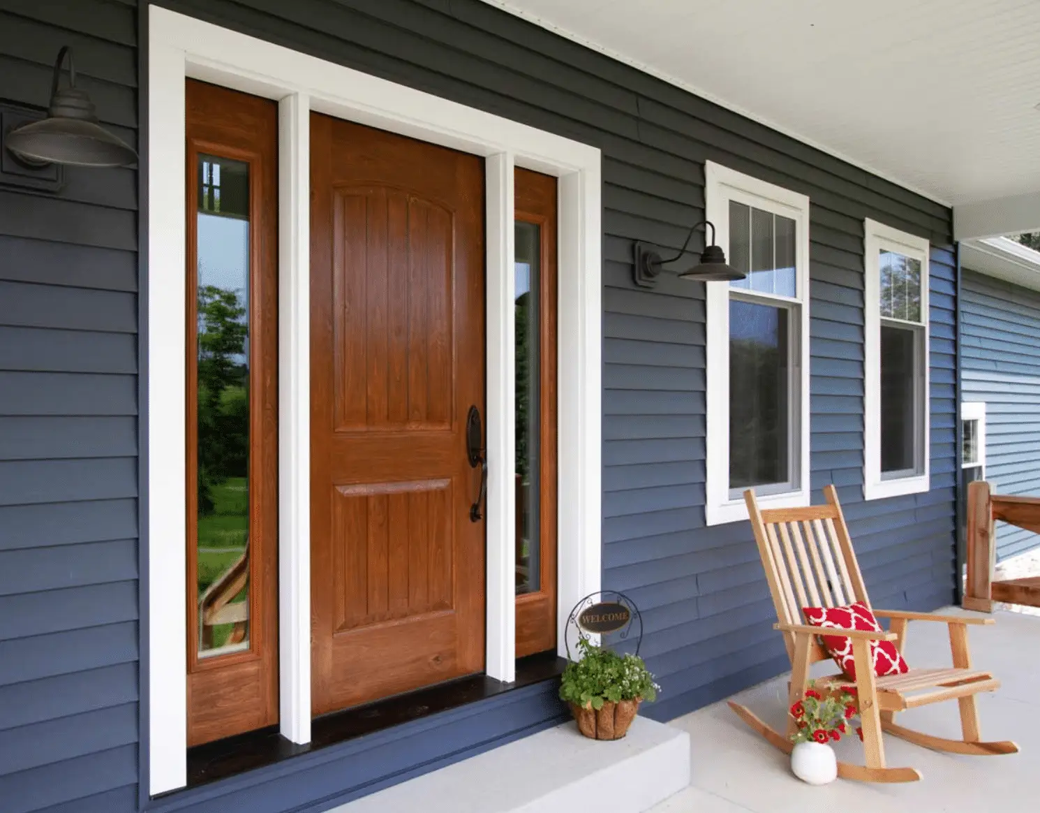 A warm knotty alder entry door with decorative sidelights complements dark blue siding and crisp white trim on a covered porch. Including the front entrance on a home maintenance checklist helps homeowners monitor the door finish, weatherstripping, threshold, locks, glass, and surrounding trim.