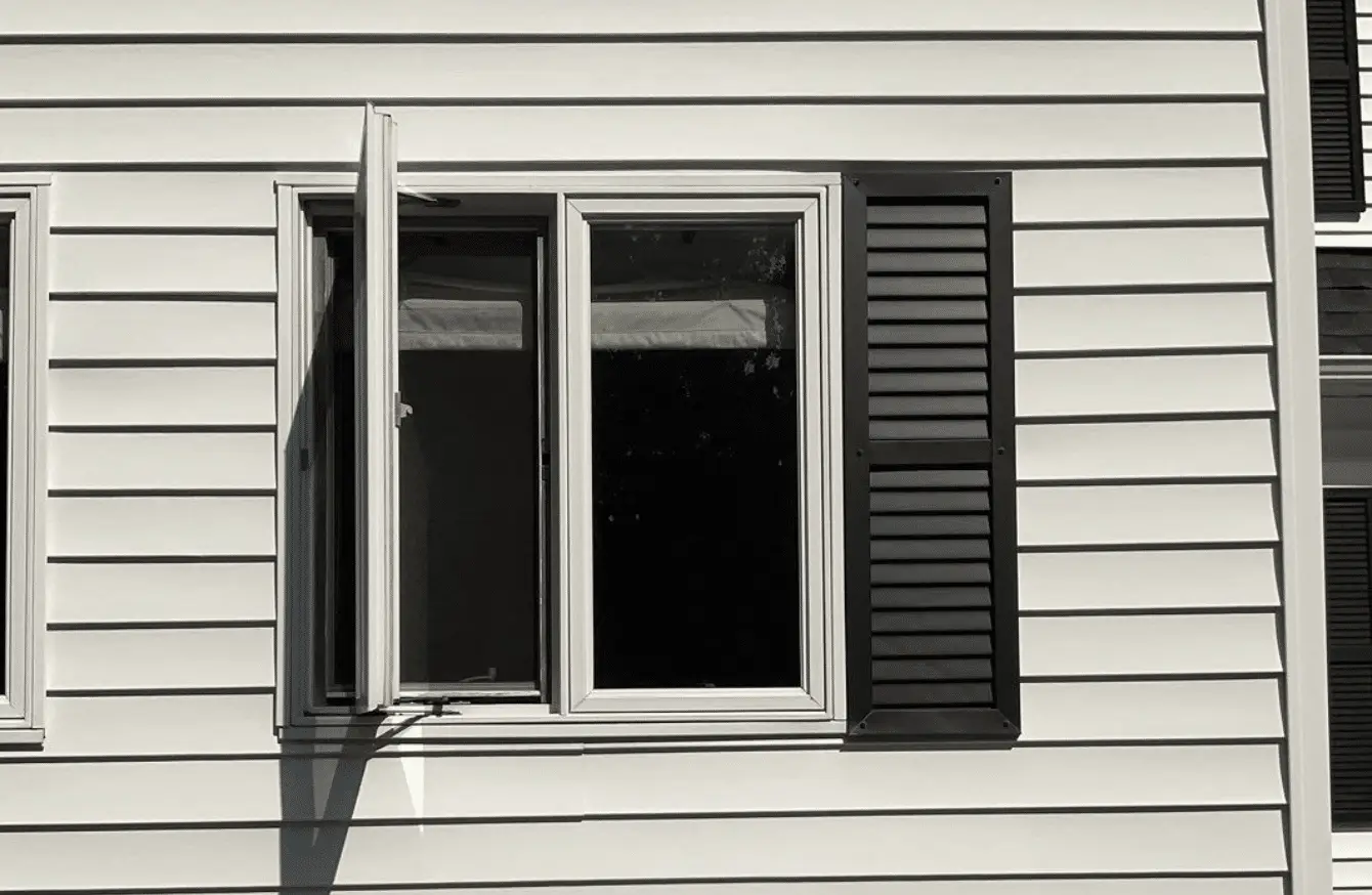 An open white basement window is surrounded by light-colored vinyl siding and black shutters on a Beverly home. A seasonal home maintenance checklist should include testing window operation, inspecting the frame, and checking for gaps that may allow drafts or moisture indoors.
