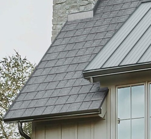 A close-up of a house roof featuring gray shingles and a section of standing seam metal roofing, with a stone chimney in the background and part of a window below the roofline.