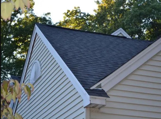 A close-up of a house with light-colored siding and a dark gray shingled roof, expertly crafted by a roofing contractor Cambridge, surrounded by green trees in the background.