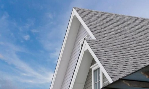 Gray shingled roof with white trim on a house, set against a blue sky with wispy clouds. A window is visible below the roof peak.