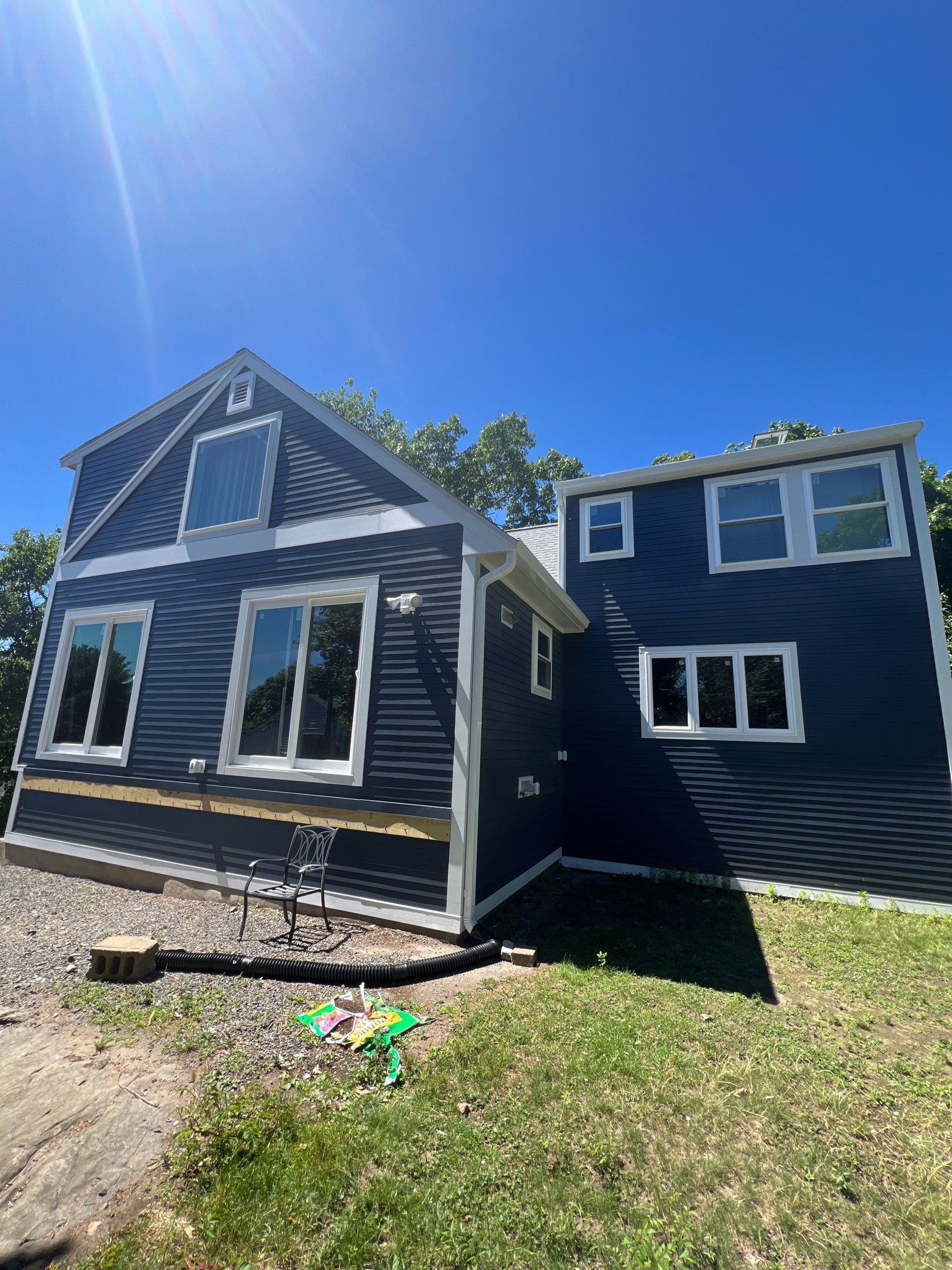 A modern blue house with white trim, large windows, and a gravel walkway in Haverhill MA. A small metal chair sits on the side, and green toys are scattered on the grass under a clear blue sky.