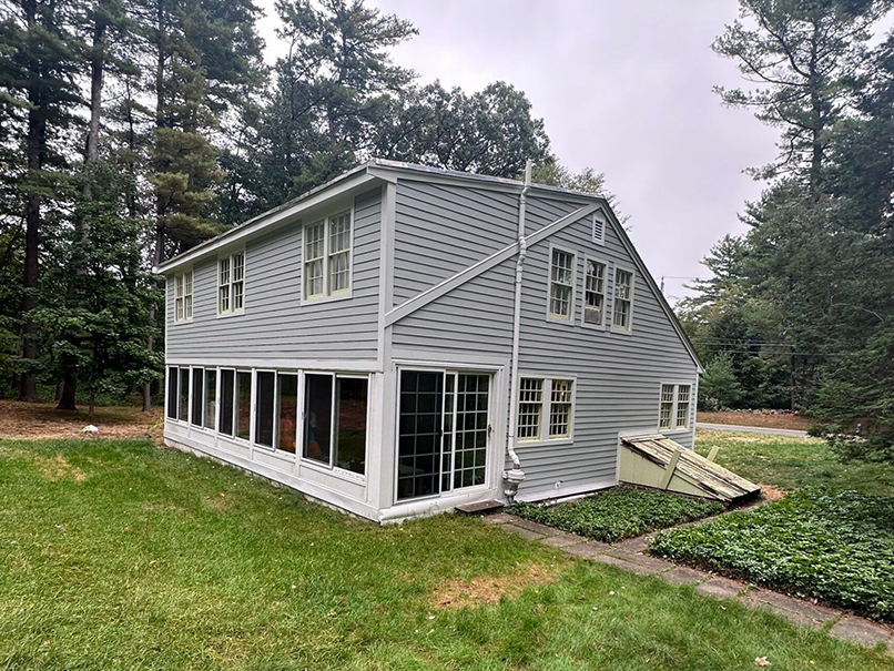 A two-story gray house with large windows and an enclosed porch sits on a grassy lawn, surrounded by tall evergreen trees on a cloudy day—an ideal project for a Siding Contractor PPC campaign.