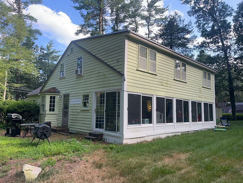 A two-story light green house with many windows and a large enclosed porch stands amid trees on a sunny day. The lawn, outdoor grills, and chair create a welcoming scene—an ideal showcase for Siding Contractor PPC campaigns.