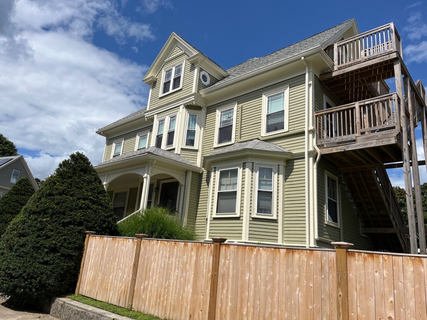 A large, light green house with white trim and multiple windows in Haverhill MA, featuring a covered porch and a tall wooden balcony and staircase on the side, behind a light wood fence and green shrubs under a blue sky with clouds.