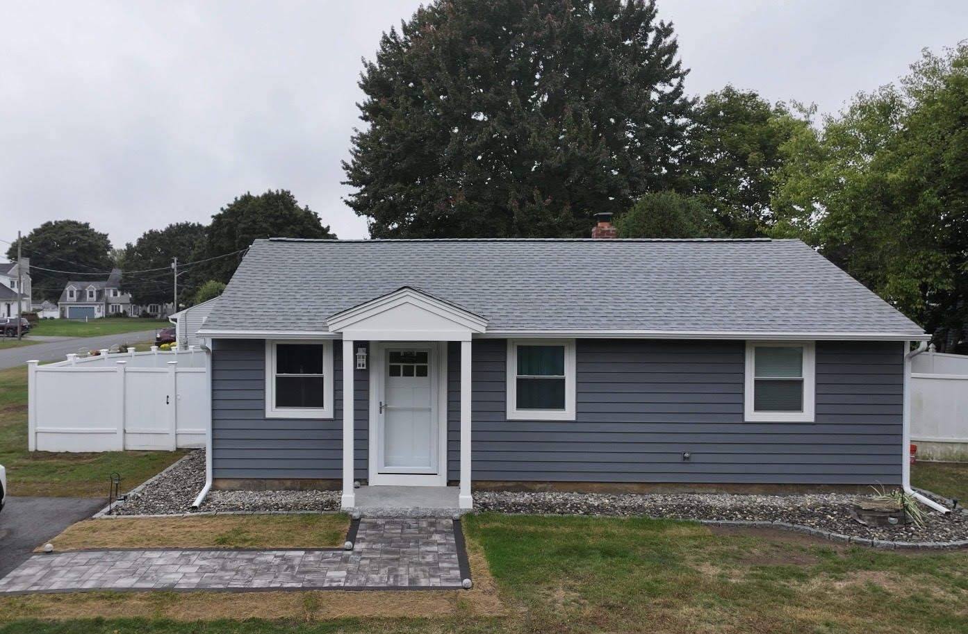 Single-story house with gray siding, white trim, and a small front porch. Perfect for installing siding in the summer, it features a paved walkway to the front door, white fencing, and patches of grass and trees in the background.