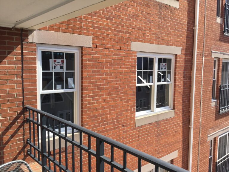 A brick building with white-trimmed Harvey Slimline windows designed for maximum glass area, some open and displaying ARVIE signs. A black metal railing runs along a balcony in the foreground.