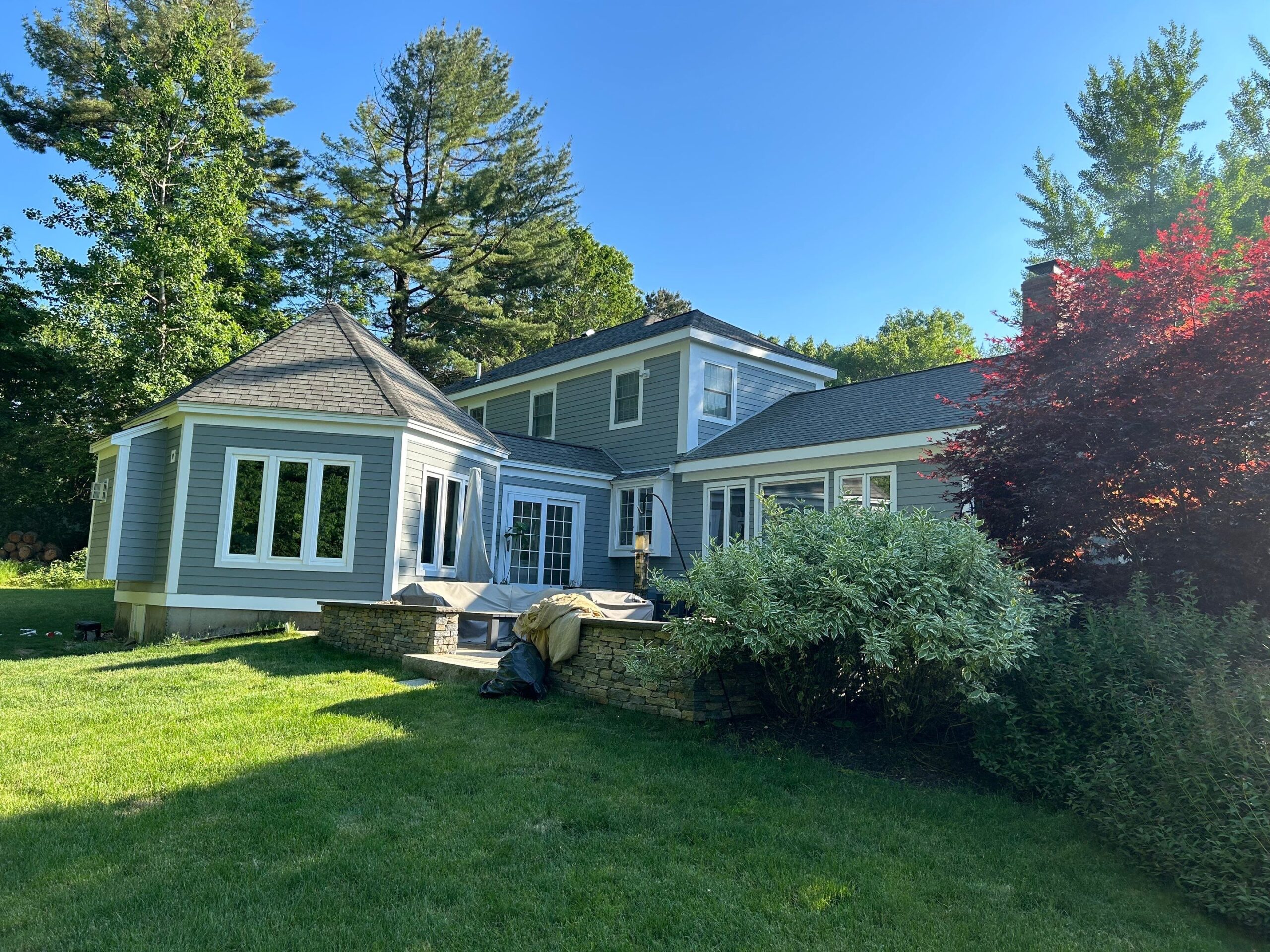 A gray two-story house with white trim sits in a lush, green yard. Large windows—perfect candidates to replace for improved energy efficiency—and a small stone patio are visible, surrounded by trees, shrubs, and a red-leafed bush under a clear blue sky.