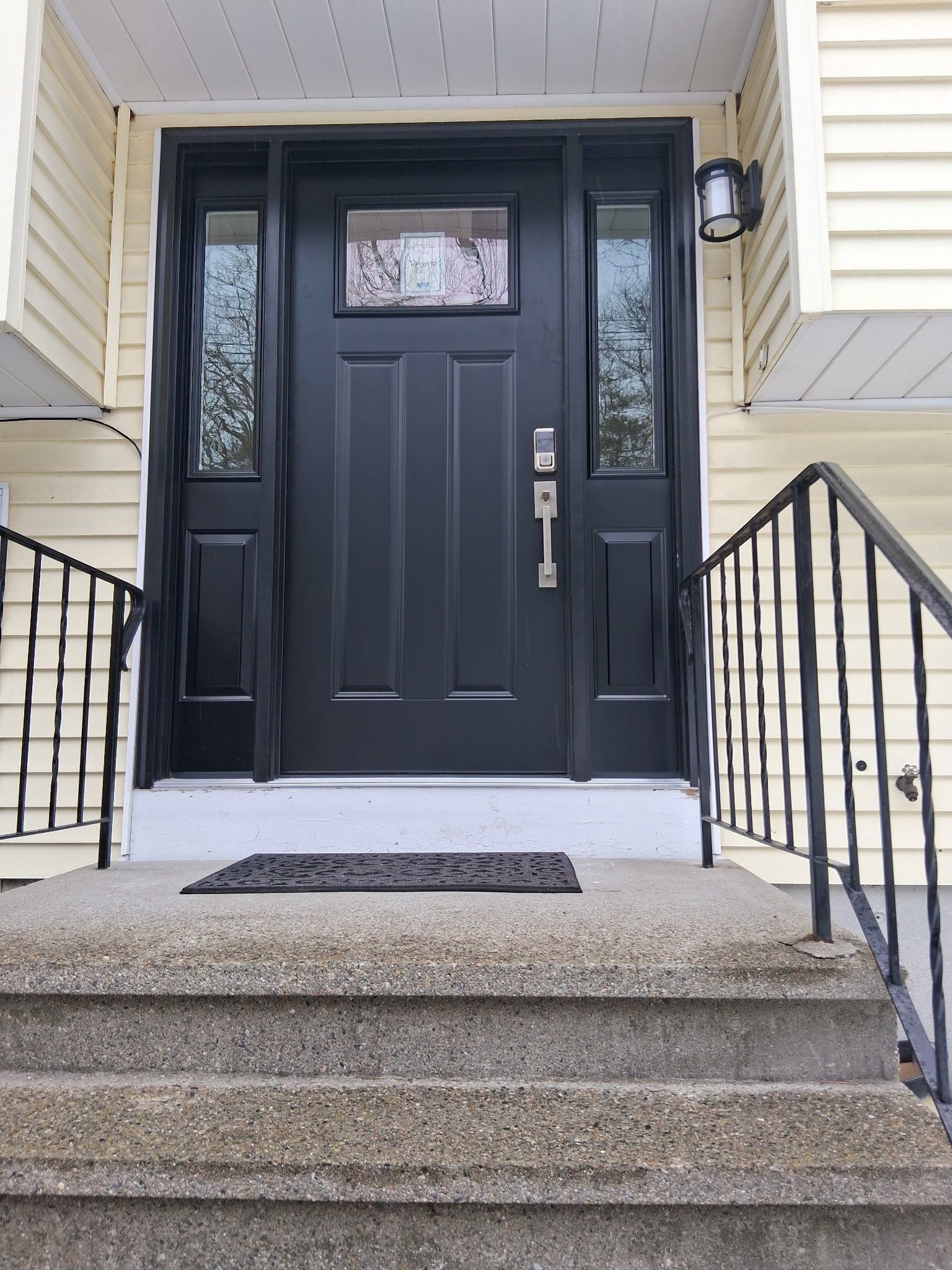 Wrought iron railings lead up concrete steps to a striking black steel entry door framed by two narrow glass sidelites against pale yellow siding. Upgrades like this define energy efficient doors in Natick MA.