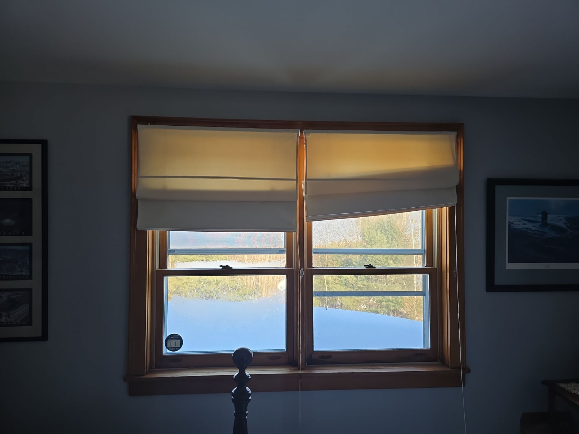 Two Roman shades are drawn halfway over a pair of older wood windows in a dim room with framed pictures on the walls. These were updated during the window installation in Ossipee.