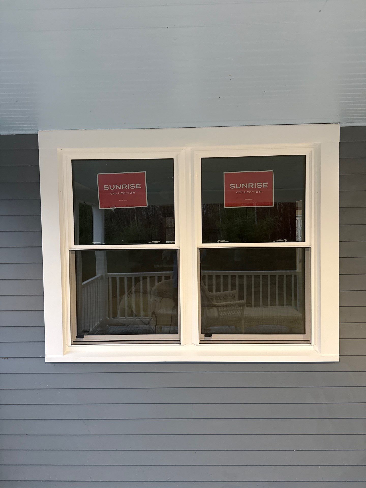 Under a porch ceiling, a pair of new white double-hung windows sits in gray siding, still showing their manufacturer labels. The fresh exterior fit shows how this window installation in Ossipee sealed out drafts.