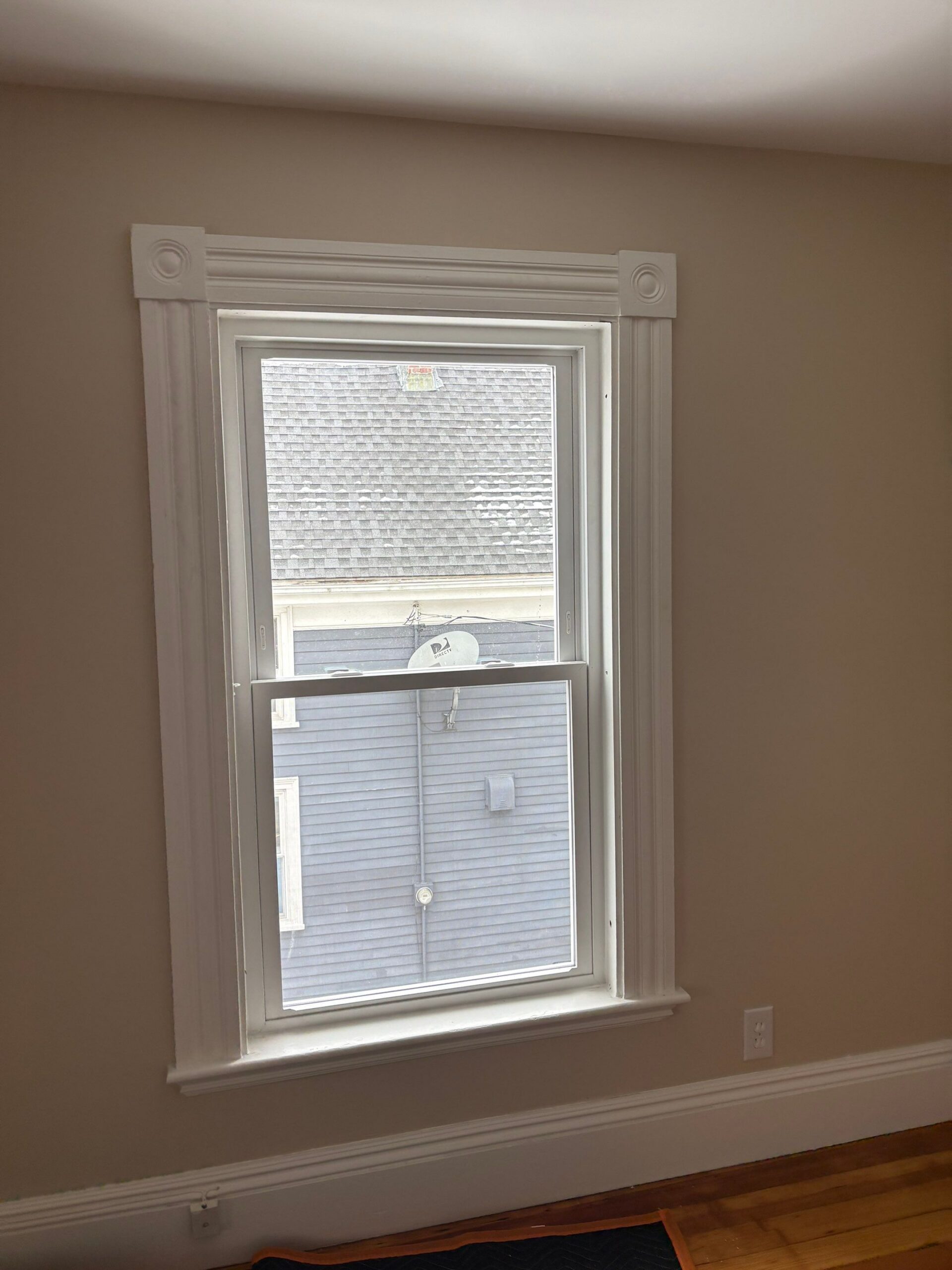 Framed by classic rosette casing, a new white double-hung lets soft daylight into a warm beige bedroom with views of neighboring rooftops. Double-pane glass gives this window installation in Salem MA steady year-round comfort.