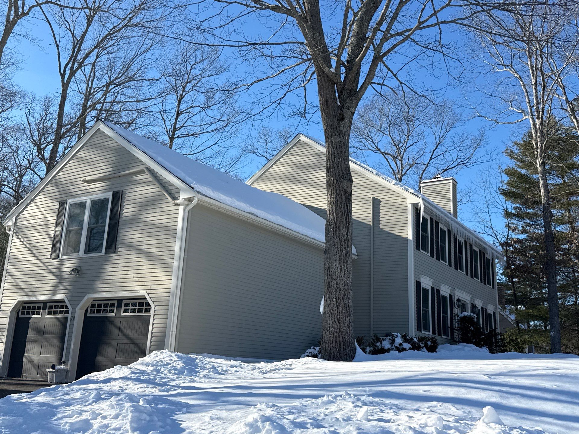 Deep snow blankets the garage and main roof of a Beverly colonial on a bright winter day, with icicles forming along the far eave. Conditions like these prompted an emergency call to a roofing contractor in Beverly MA for ice dam help.