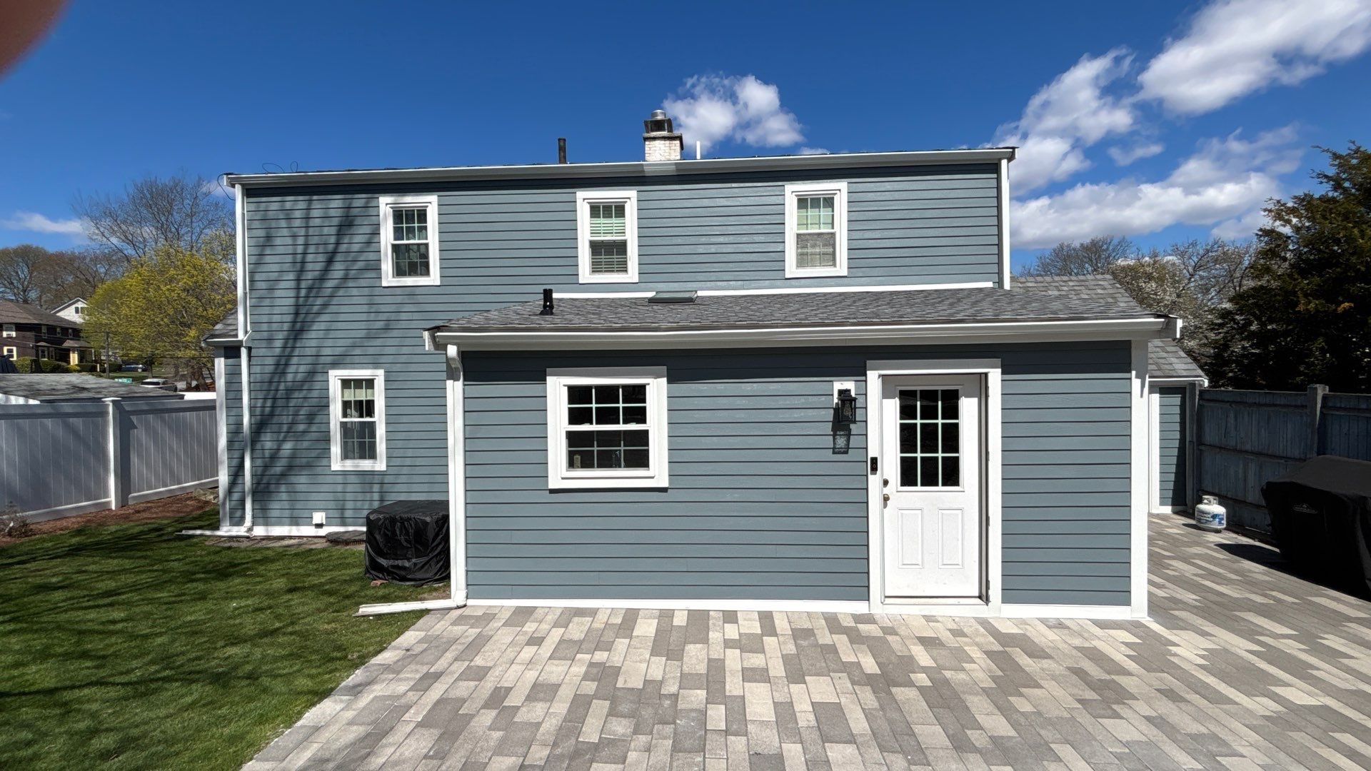 Looking straight at the back, Boothbay Blue lap siding covers both stories and the single-story addition above a paver patio. Bright white casings around every window and door mark the finish from James Hardie certified installers in Wakefield, MA.