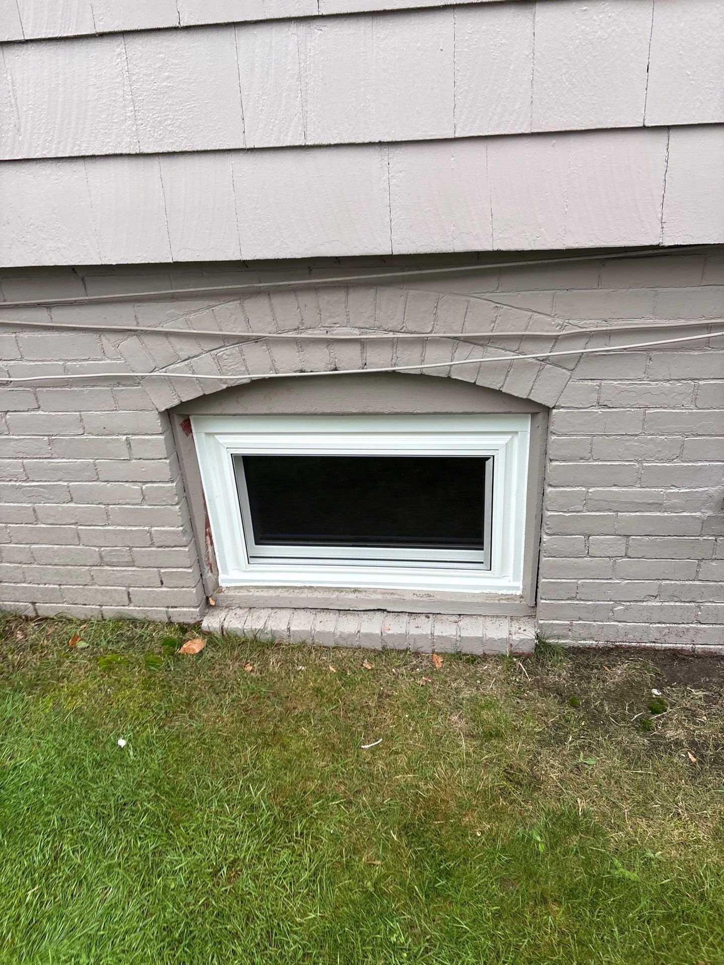 Close up, a bright white basement window sits squarely under a painted arched brick header and gray cedar shingles above a strip of lawn. The tight fit shows the craftsmanship behind this window installation in Winthrop.