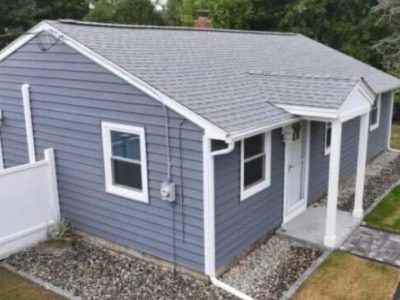 A small, single-story house with blue-gray vinyl siding in Carlisle MA, white trim, and a shingled roof. There are two windows on the visible side and a white door with a small covered entryway. The yard features gravel borders and some grass.