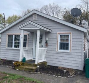 A small gray house with white trim and vinyl siding in Arlington MA features a front porch, two windows, and a dish antenna on the roof. Flower beds, a green trash bin, and a dumpster with a sign reading CAMS sit beside the house.