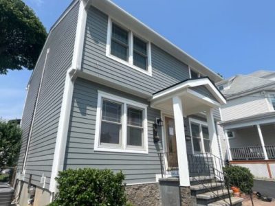 A two-story house with gray vinyl siding in Carlisle MA, white trim, and a small covered front entrance. The house features double-pane windows, a stone foundation, and some greenery near the front steps under a clear blue sky.