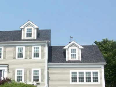 A two-story gray house with white trim and shutters, featuring multiple windows and two small dormers on the roof, showcases the quality work of roofing contractors in Bedford MA, surrounded by greenery and set against a clear blue sky.