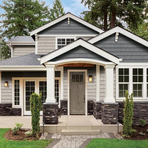 A charming gray house with white trim, stone accents, and a covered front porch features quality vinyl siding in Rowley MA, surrounded by green grass, shrubs, and tall trees in the background.