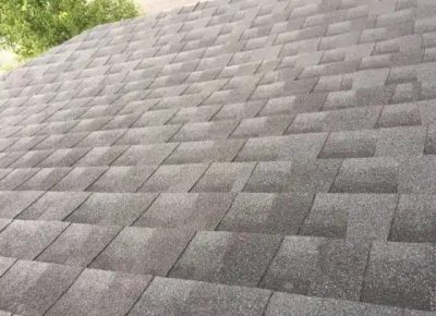A close-up view of an asphalt shingle roof by a trusted Roofing Company in Chestnut Hill, with overlapping rectangular shingles in varying shades of gray. The offset pattern and green tree leaves add visual interest in the upper left corner.