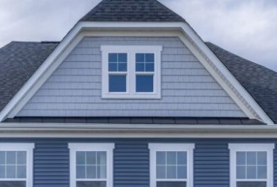 The image shows the upper section of a house with gray shingles, blue vinyl siding in Belmont MA, three sets of white-trimmed windows, and a steep gable roof under a cloudy sky.