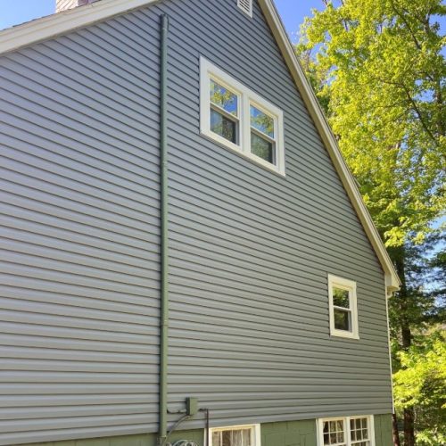 A gray house with white trim on the windows, vinyl siding in Rowley MA, features a green hose reel by the foundation and is surrounded by tall trees under a clear blue sky.