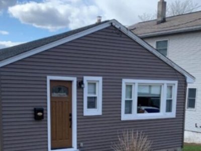 A small single-story house with brown siding, Replacement Windows in Arlington MA with white trim, a wooden front door, a small set of steps, and a brown shrub in the front yard. Another taller house is visible in the background.